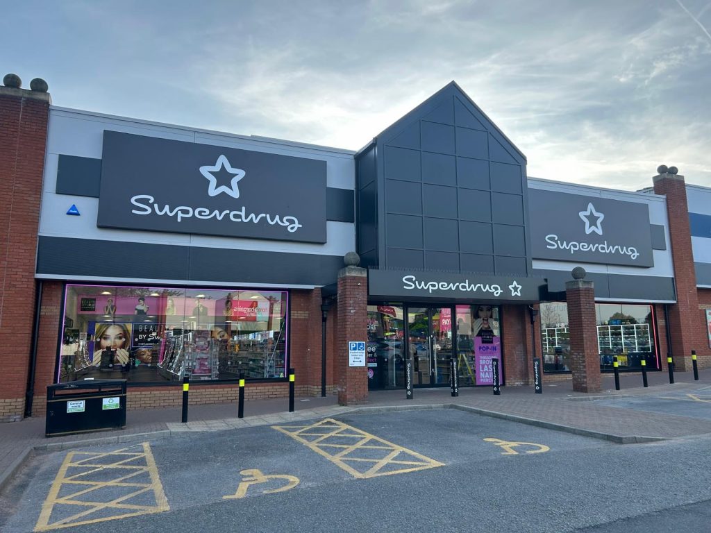 Exterior of Superdrug Crewe store featuring illuminated signage, branded tray signs, and pink-accented shopfront windows with promotional displays.